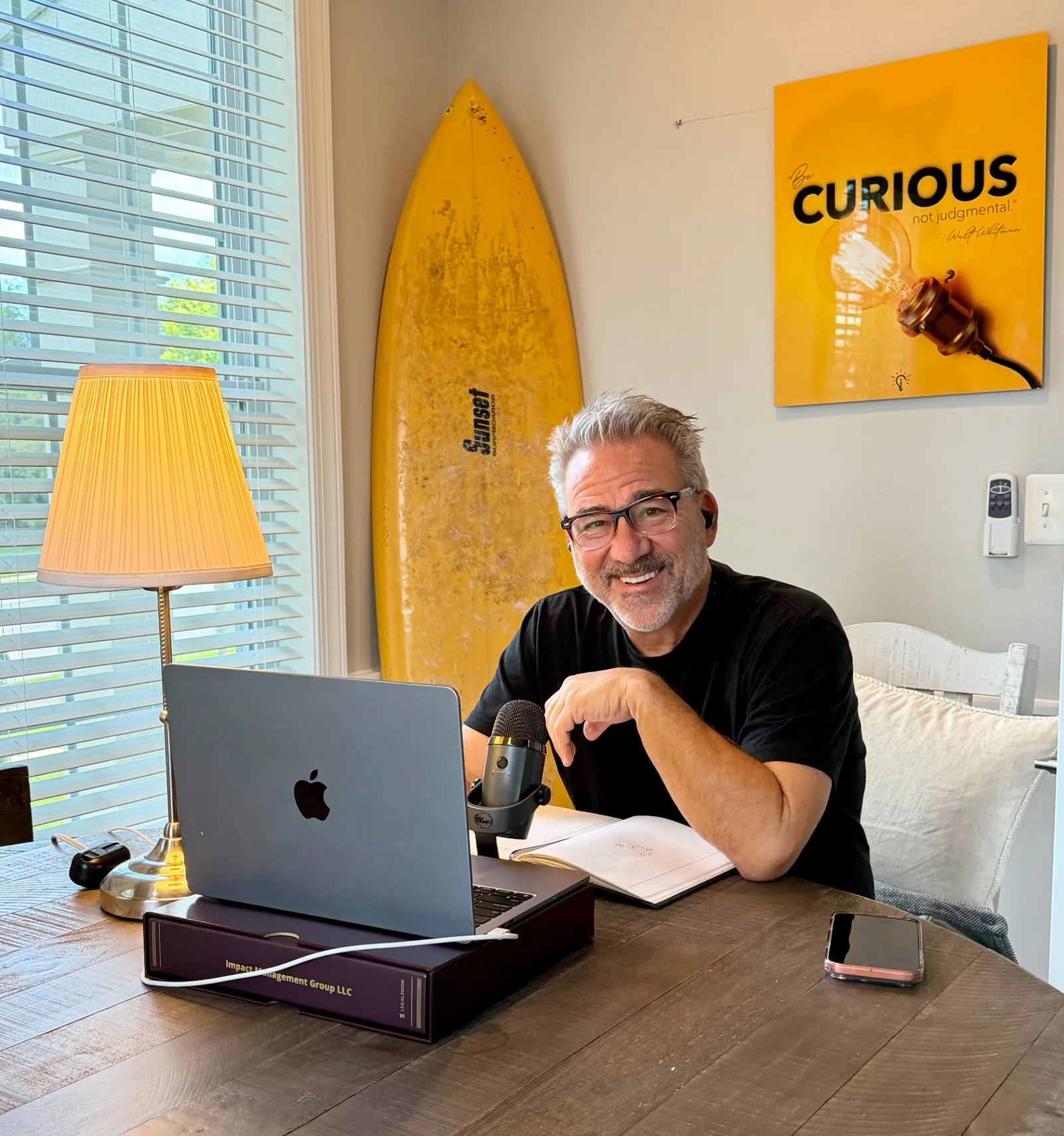 Mark P. Fisher smiling at the camera from his conversation room with a microphone, laptop, surfboard, and Curious sign